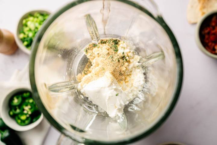 A blender filled with cream cheese, chopped garlic, and herbs, viewed from above. Surrounding bowls hold sliced jalapeños and spices.